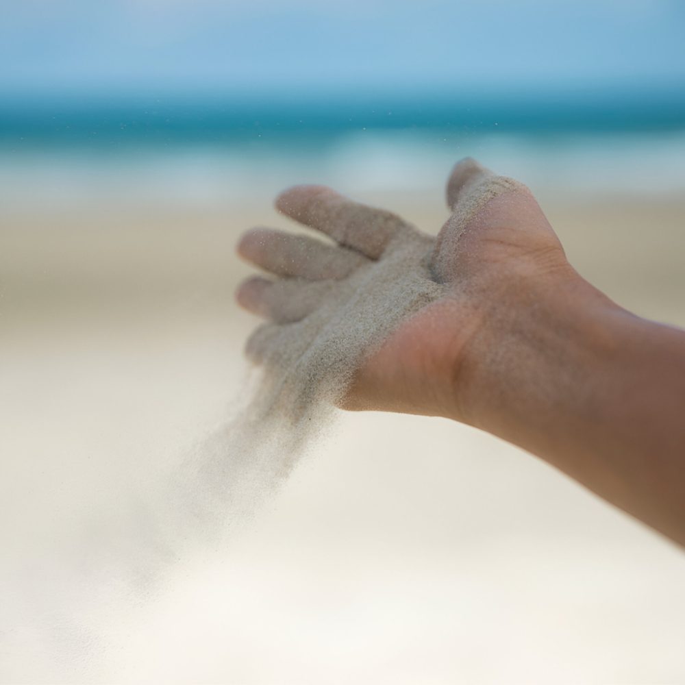 Lâcher-prise : le sable qui s’écoule entre les doigts alt = "Main laissant s’écouler du sable sur une plage avec l’océan en arrière-plan, symbole du lâcher-prise et du temps qui passe"